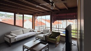 A living room with a sofa, armchair, coffee table, and TV, with large windows offering a view of buildings and a red-roofed area. - Nirvana Otel  (Bozcaada)
