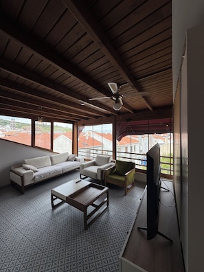A living room with a sofa, armchair, coffee table, and TV, with large windows offering a view of buildings and a red-roofed area.