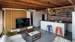 A modern living room with a TV, sofa, coffee table, and kitchen area with stools. - Nirvana Otel  (Bozcaada)