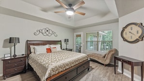 A bedroom with a bed, bedside tables, a chair, a desk, a ceiling fan, and a large wall clock.