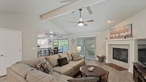 A living room with a sectional sofa, a fireplace, a ceiling fan, and a kitchen in the background.