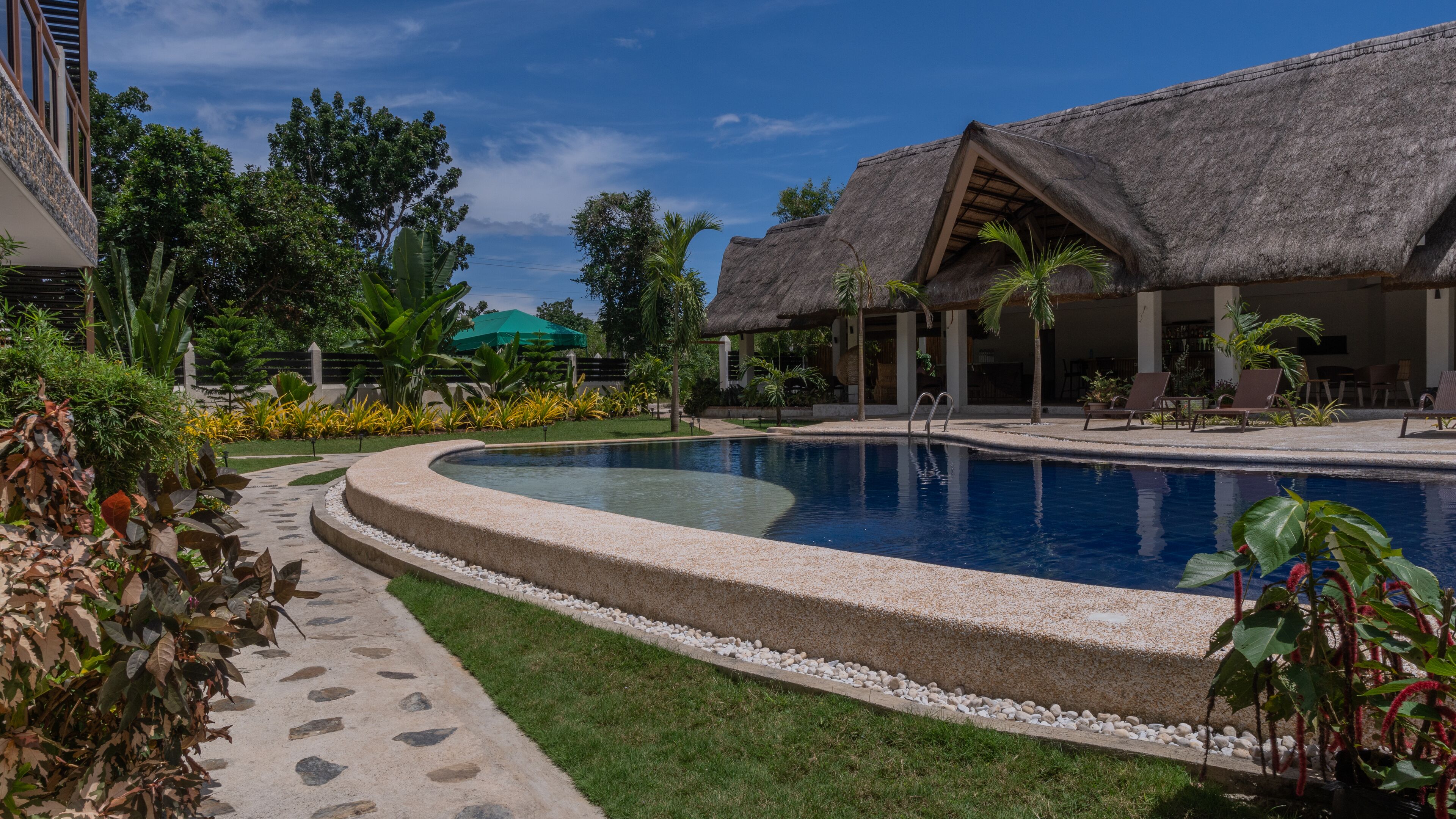 Zona de piscina en un resort, con un edificio de techo de paja, un sendero pavimentado y abundante vegetación.