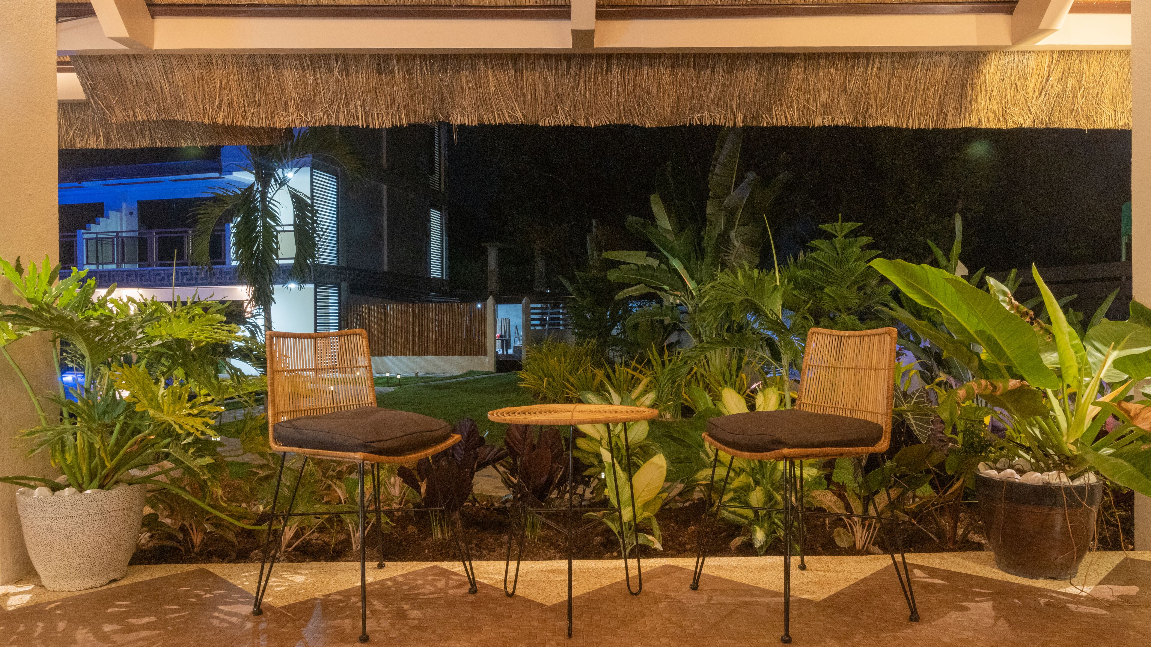 An outdoor seating area with wicker chairs and a small table, surrounded by potted plants and a thatched roof overhead.