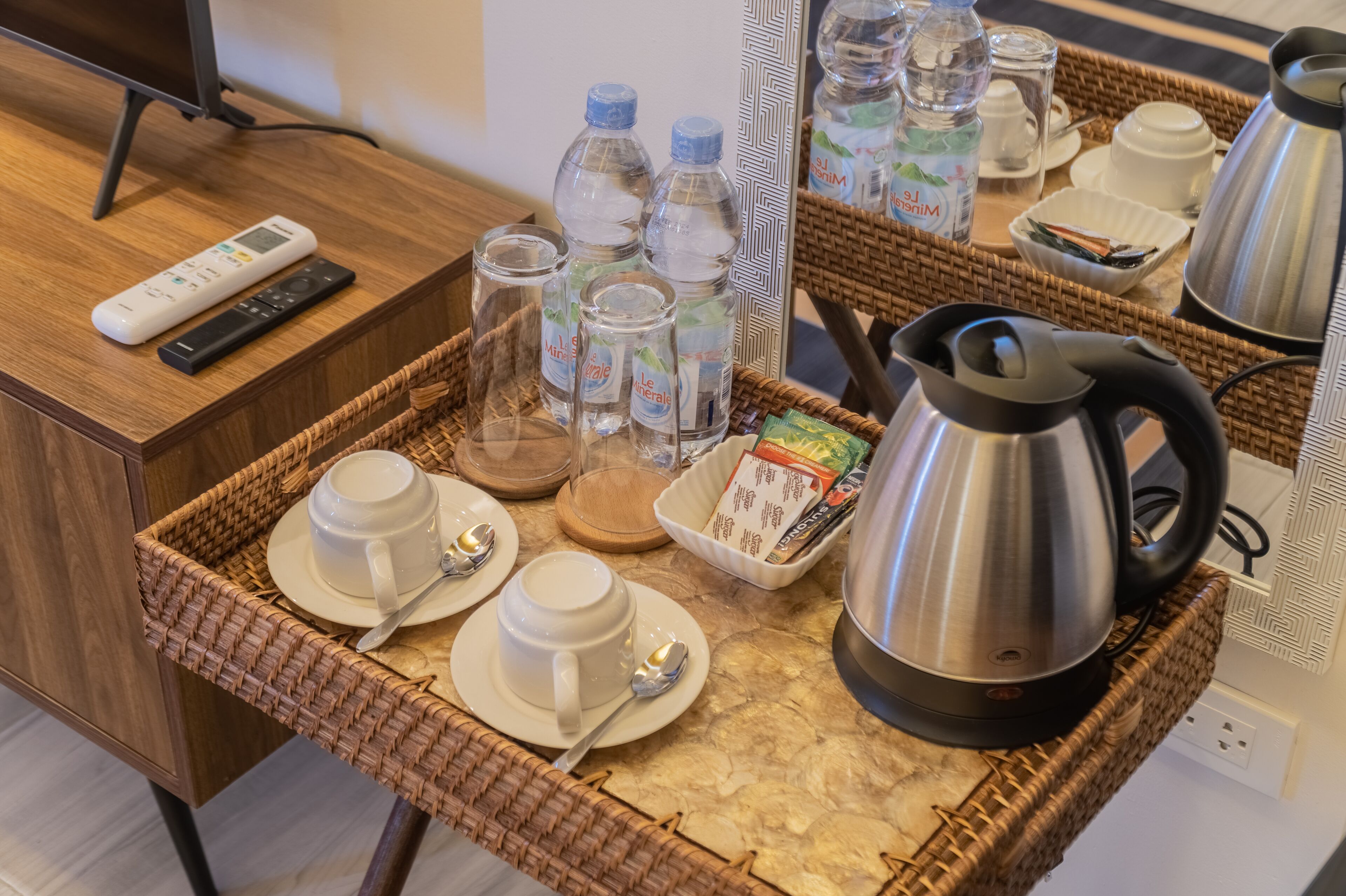 Ein Frühstücksbrett mit Tassen, Löffeln, Wasserflaschen, einem Wasserkocher und einer Fernbedienung auf einem Holztisch.