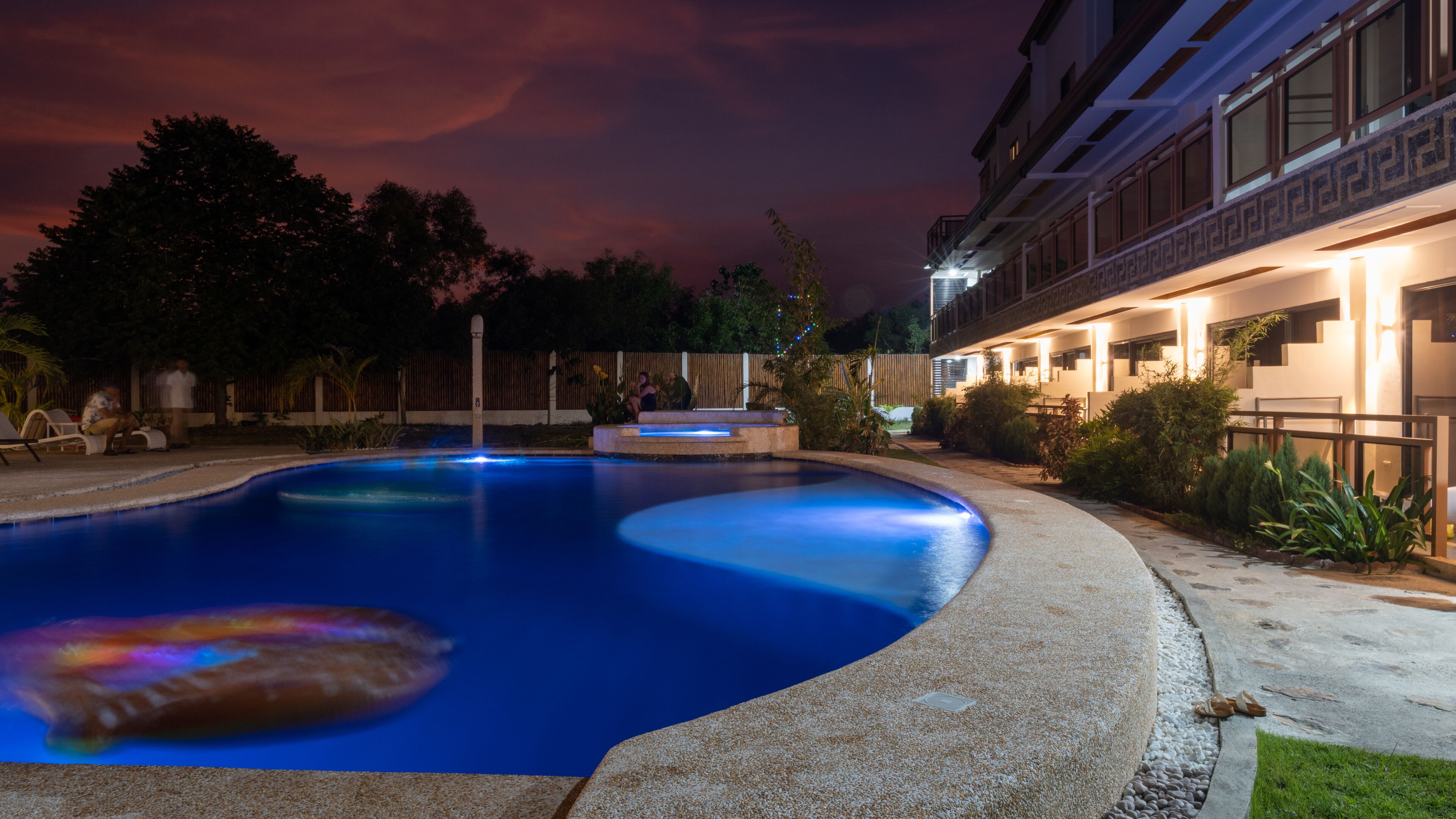 Une piscine avec un jacuzzi, entourée d’une clôture et d’arbres. On aperçoit, au fond, un bâtiment dont les fenêtres sont illuminées.