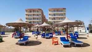 A beachside resort with multiple lounge chairs and umbrellas, a pool area, and a multi-story building in the background. - Studio flat with balcony, free access to beach and pools at Casablanca Beach (Hurghada)