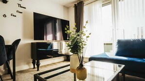 Un salon avec un téléviseur à écran plat, un canapé bleu foncé, une table basse en verre et une plante.