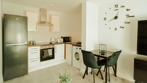 Une cuisine moderne équipée d’un réfrigérateur, d’un four et d’un four à micro-ondes en acier inoxydable. On y trouve un coin repas avec deux chaises et une table. Une horloge est fixée au mur.