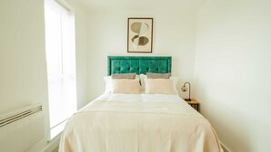 Une chambre à coucher avec une tête de lit recouverte d’un tissu vert, une couvre-lit blanche et une œuvre d’art abstraite encadrée au-dessus du lit.
