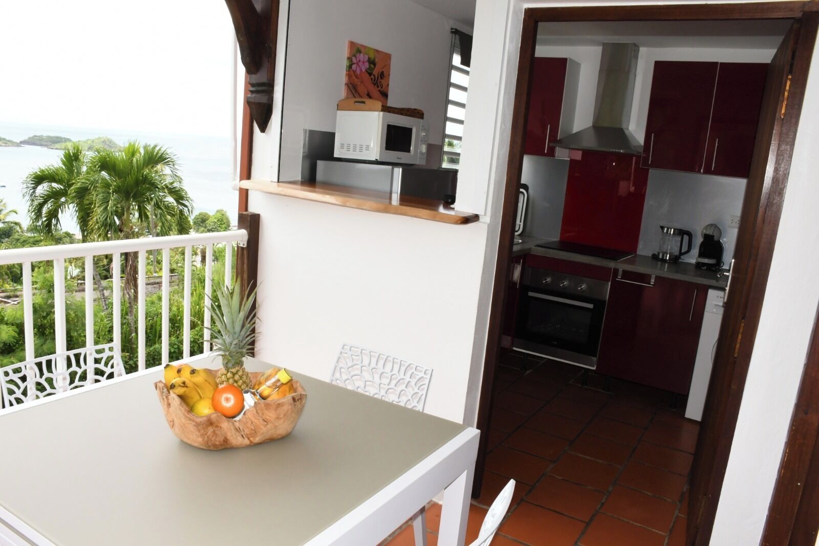 Un balcon avec une table sur laquelle se trouvent un panier de fruits, un four à micro-ondes et un tableau. On aperçoit la cuisine, avec ses meubles rouges et sa cuisinière.