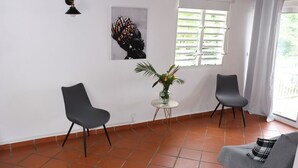 Una habitación con dos sillas, una mesita con un florero, un cuadro en la pared y una ventana con persianas.