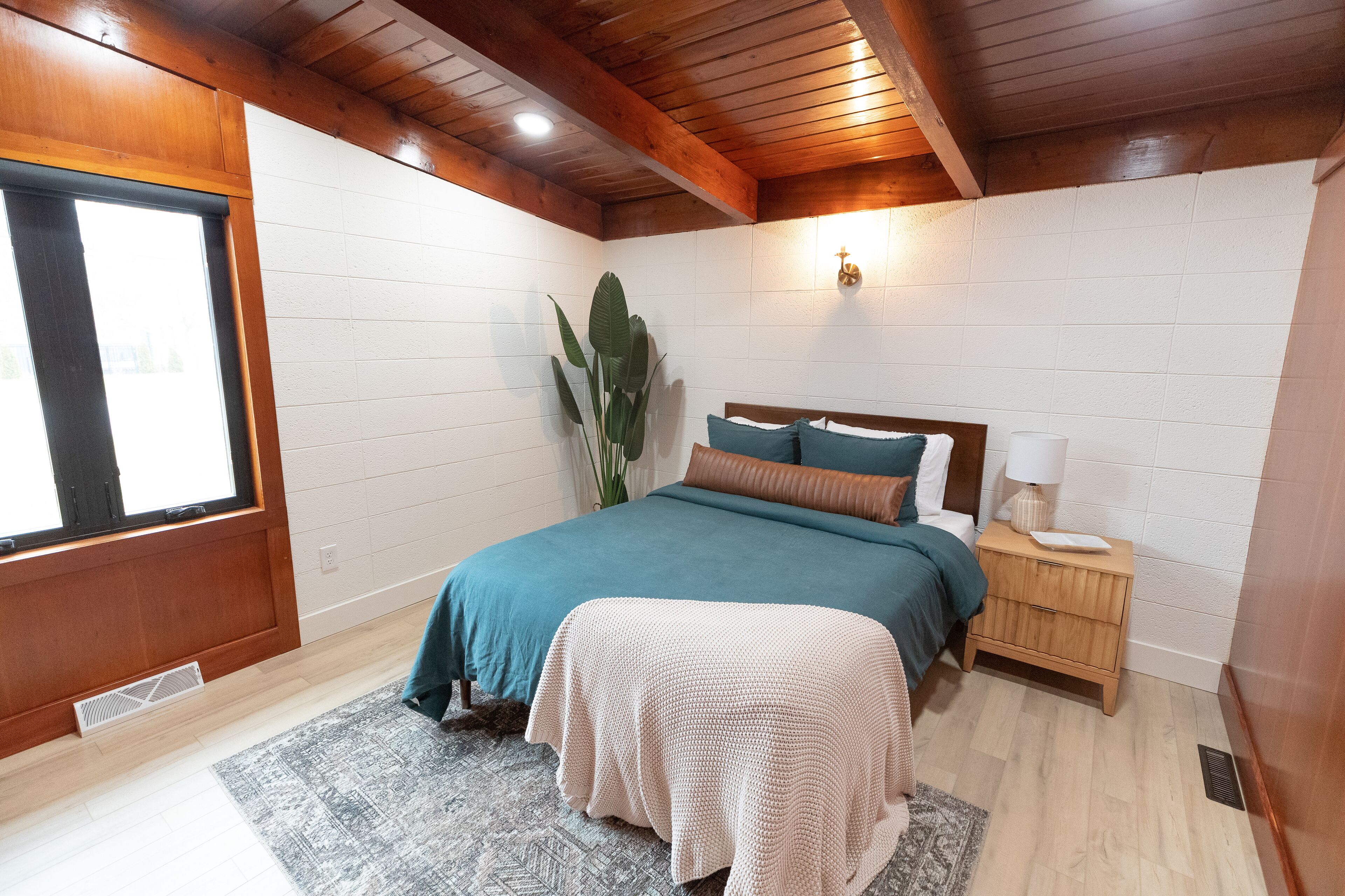 A bedroom with a bed, bedside table, lamp, and a large plant.