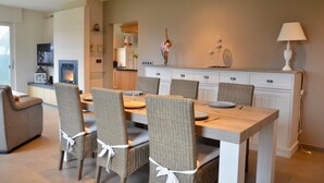 A dining area with a long table and six chairs, a fireplace, and a kitchen in the background.