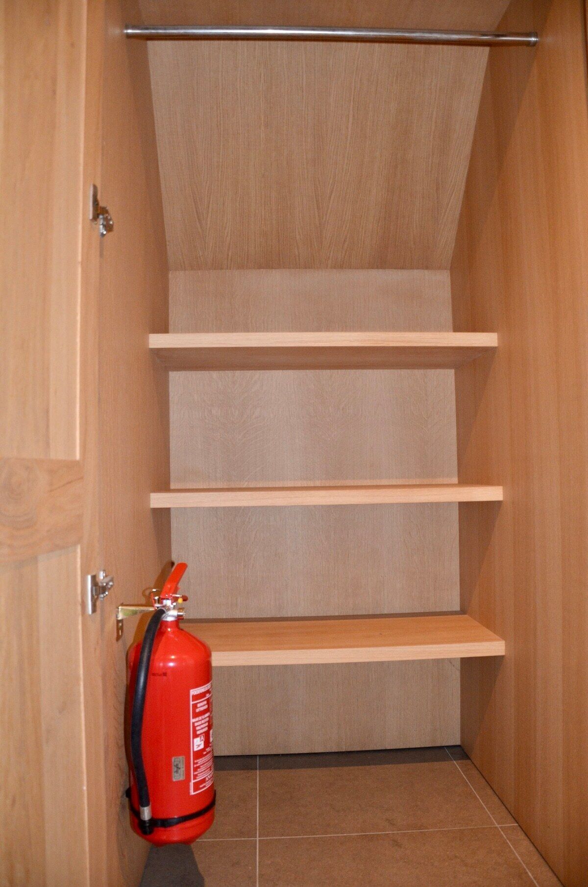 Un espacio pequeño y cerrado con paredes de madera y suelo de baldosas, que cuenta con tres estantes de madera y un extintor rojo en el suelo.