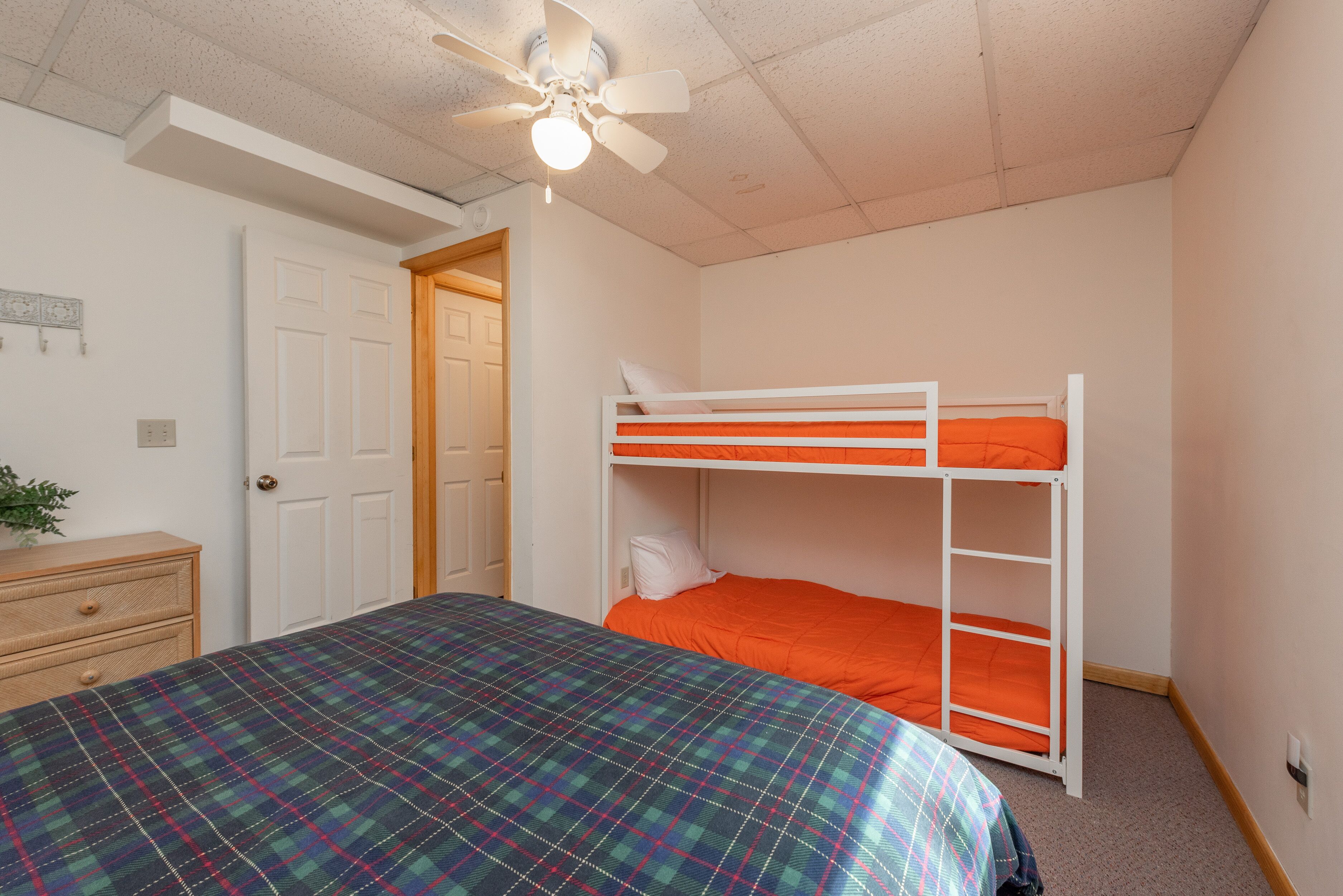 Une chambre avec un lit superposé, un lit simple recouvert d’une couverture à carreaux, une commode en bois et un ventilateur de plafond.