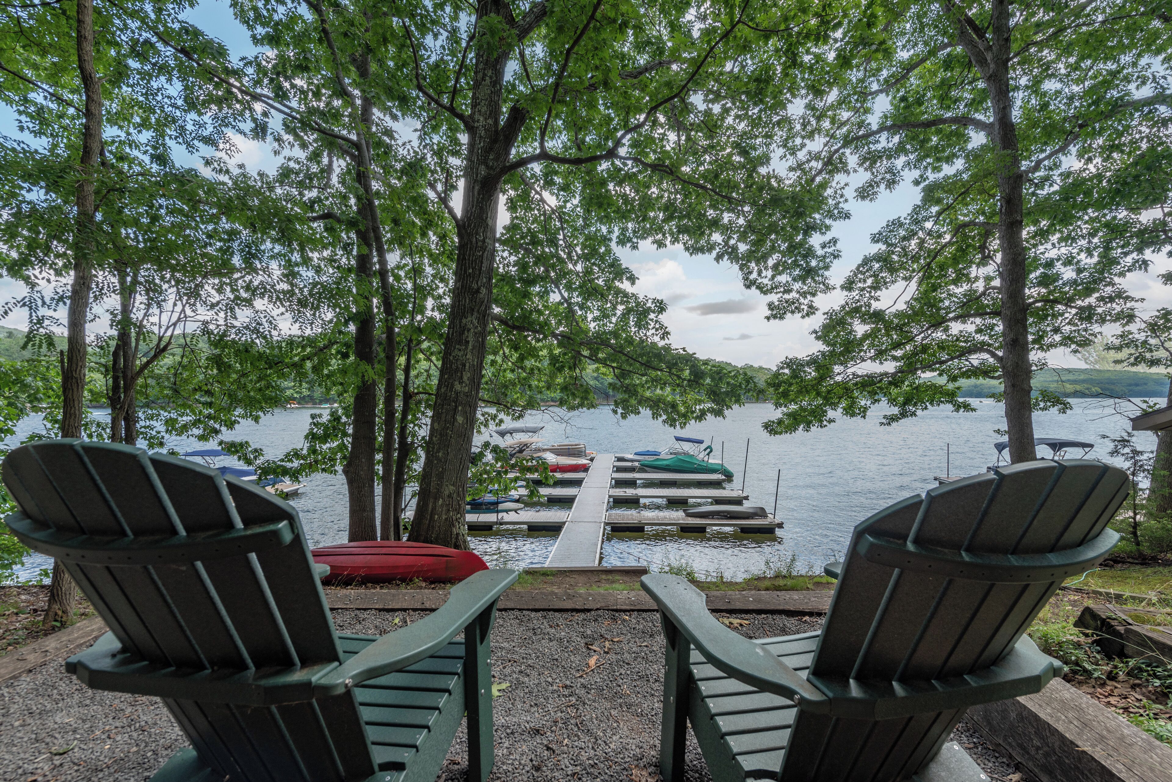 Two Adirondack chairs facing a dock with boats, surrounded by trees and a lake.