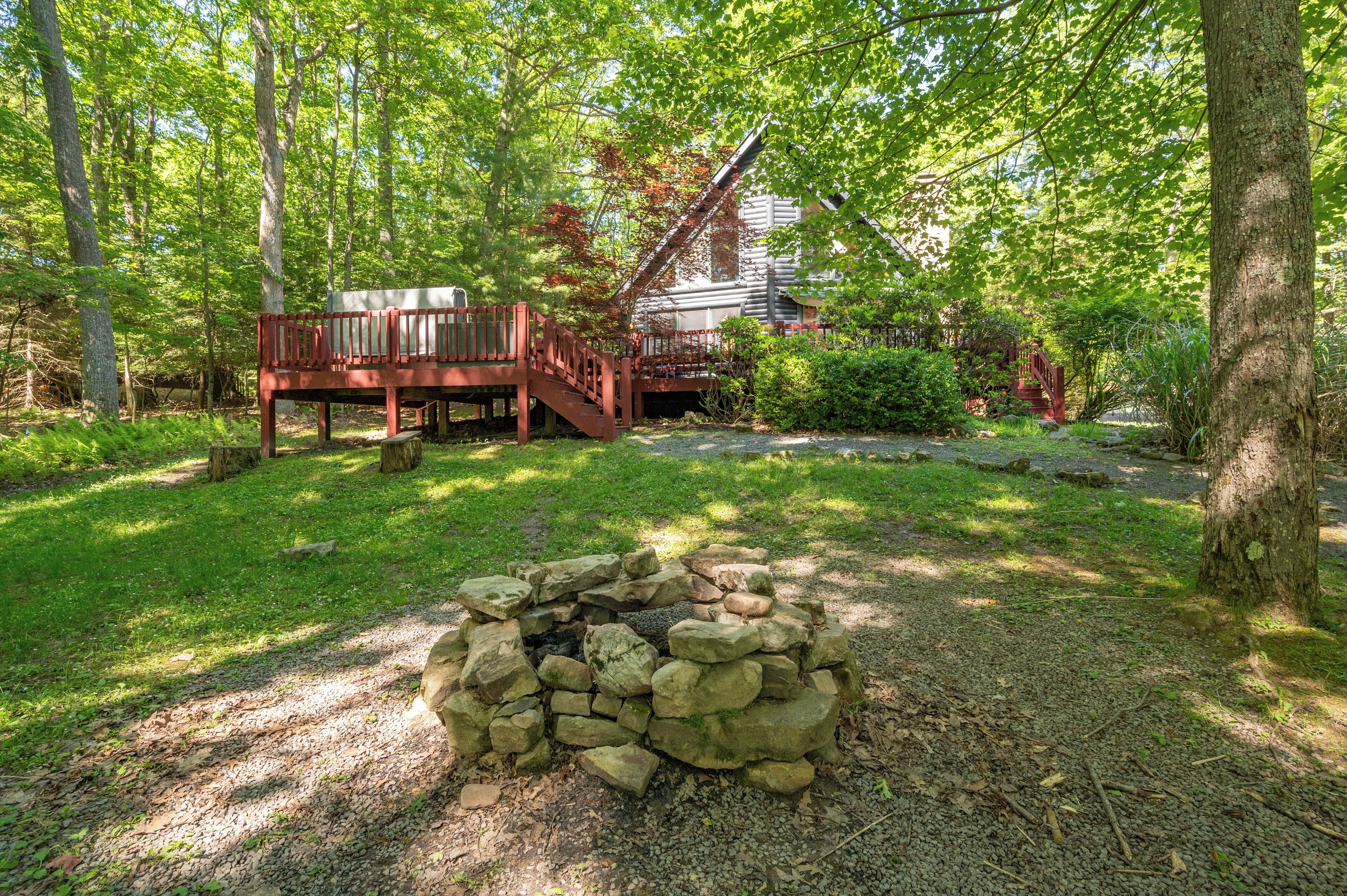 A rustic cabin with a stone fire pit in the foreground, surrounded by lush greenery and a wooden deck.
