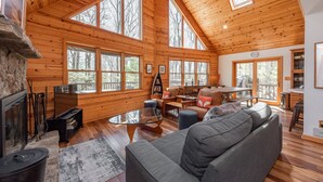 Eine geräumige Hütte mit Holzwänden und hoher Decke, mit Kamin, einer Couch, einem Esstisch und großen Fenstern mit Blick auf den umliegenden Wald.