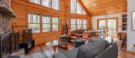 Una cabaña amplia con paredes de madera y techo alto, que cuenta con chimenea, sofá, mesa de comedor y grandes ventanales con vistas al bosque circundante.