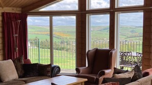 Una habitación acogedora con techo de madera, ventanas amplias y vistas a colinas verdes. Hay un sofá, una mesa y sillas.