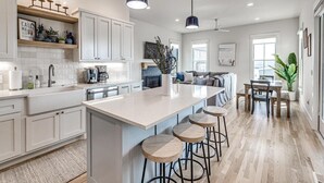 Moderni keittiö, jossa on suuri keittiösaareke, baarituolit, riippuvalaisimet sekä ruokailutila, jossa on pöytä ja tuolit.