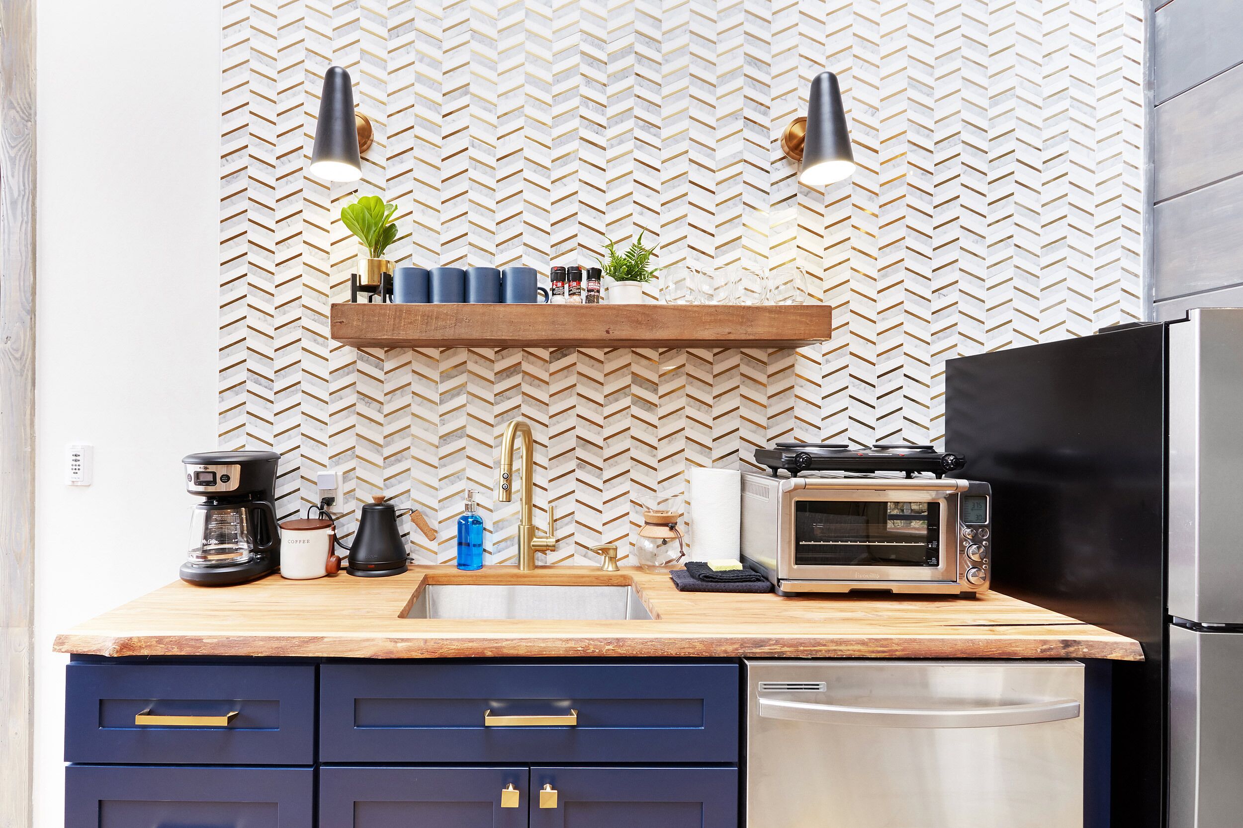 A kitchen with a herringbone-patterned backsplash, a wooden countertop, a built-in oven, a stainless steel dishwasher, and a hanging shelf with plants and kitchen items.