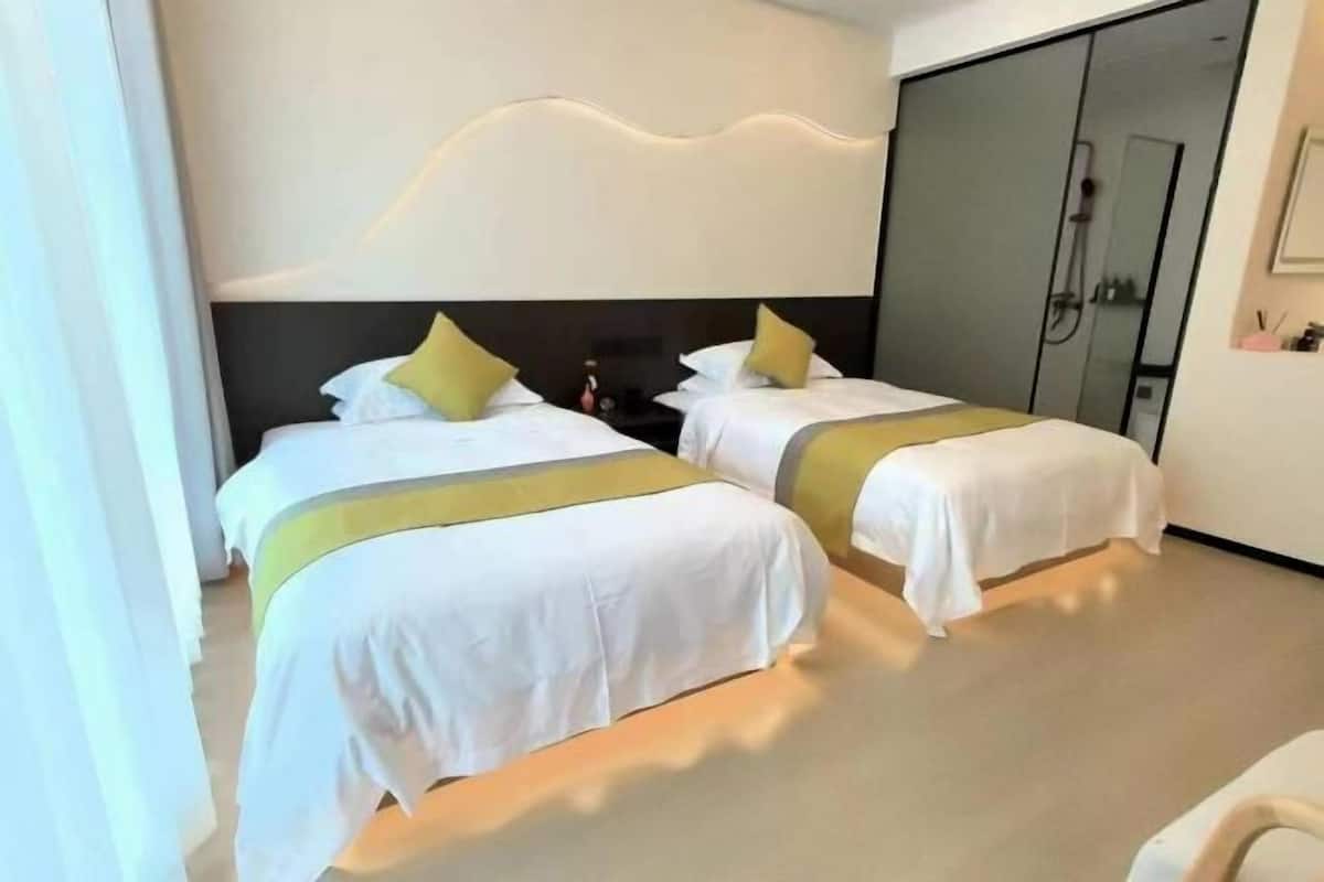 Hotellihuone, jossa on kaksi sänkyä. Jokaisessa sängyssä on valkoiset vuodevaatteet ja keltaisia yksityiskohtia, moderni pääty sekä liukuovi, joka johtaa suihkutilaan.