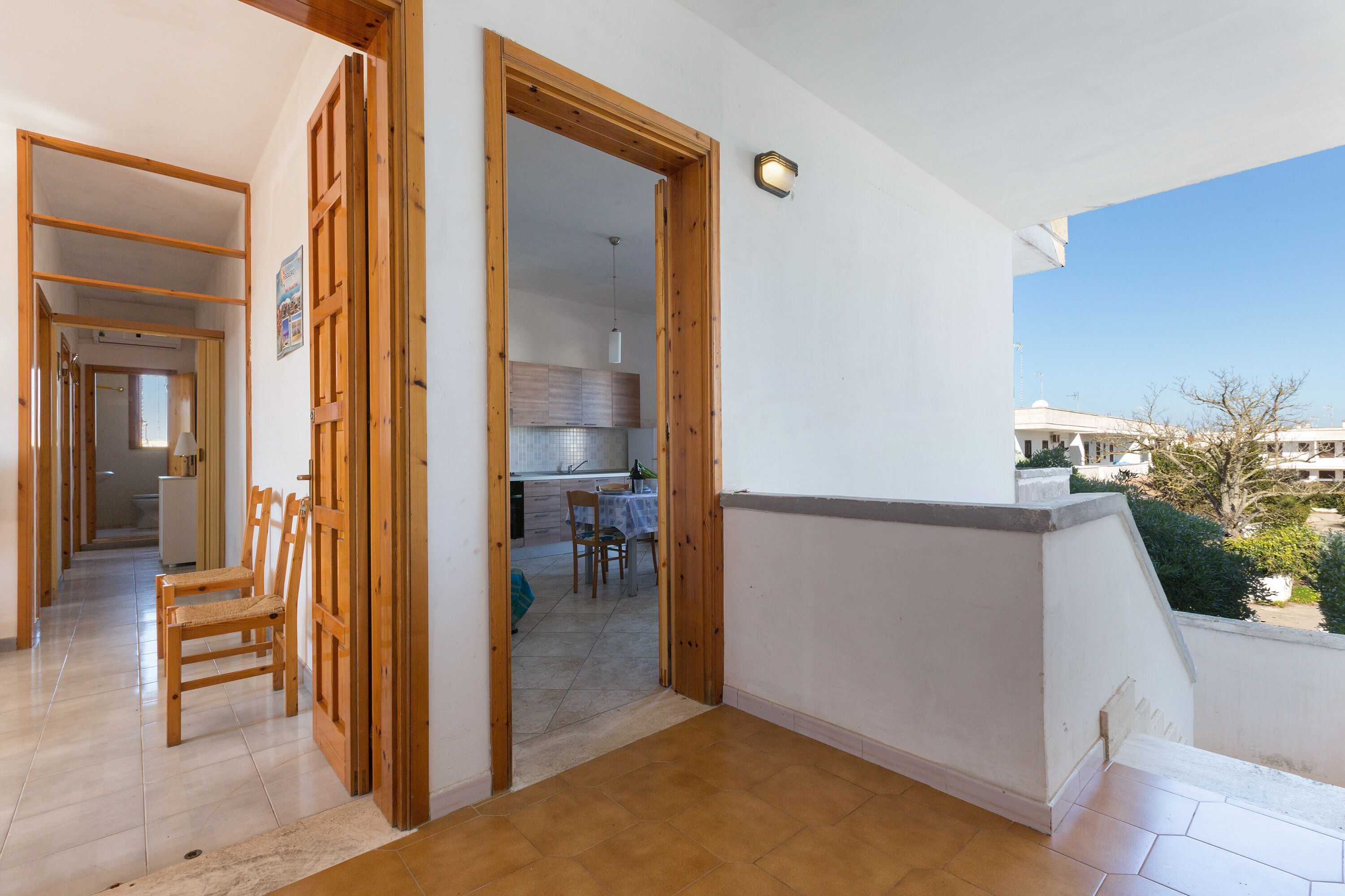 A balcony with a wooden floor, a white wall, and a view of a building and trees.
