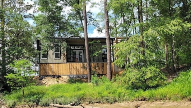 A cabin with a wooden exterior, surrounded by dense greenery and a body of water.