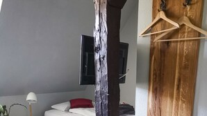 Un dormitorio con techo inclinado, una cama con ropa blanca, un cojín rojo, una mesita de noche pequeña con una lámpara y un televisor montado en la pared.