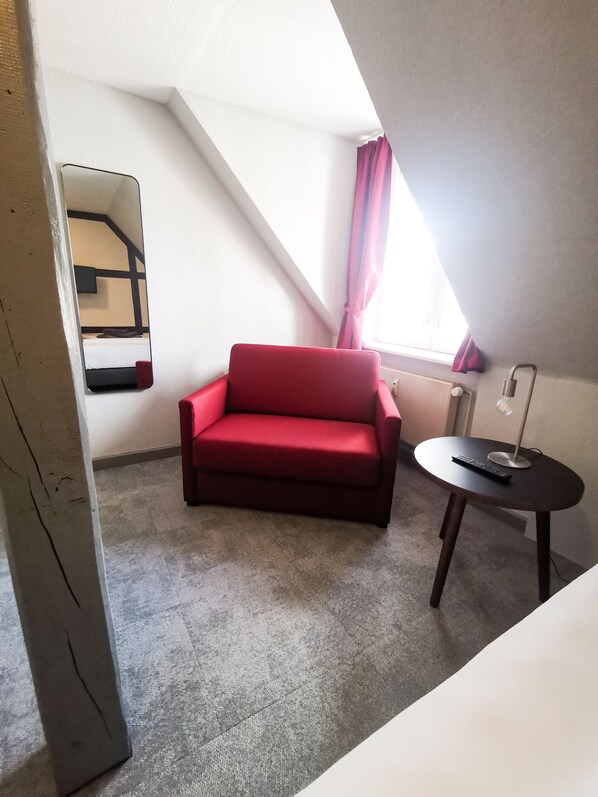 A small, red sofa in a corner of a room with a sloped ceiling, a small table with a lamp, and a window with curtains. - Kontraste (Neumuenster)