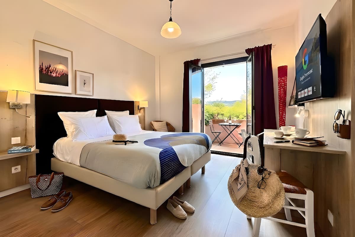 A hotel room with a bed, bedside table, lamp, and a view of the outdoors.