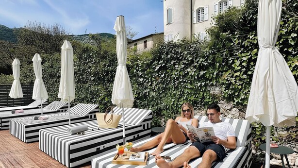 A poolside area with striped lounge chairs and umbrellas, a wooden deck, and a building with a tower in the background. - Hotel Le Provence - Restaurant Le Styx (La Palud-sur-Verdon)