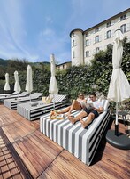 Área da piscina com espreguiçadeiras listradas e guarda-sóis, um deck de madeira e um edifício com uma torre ao fundo.