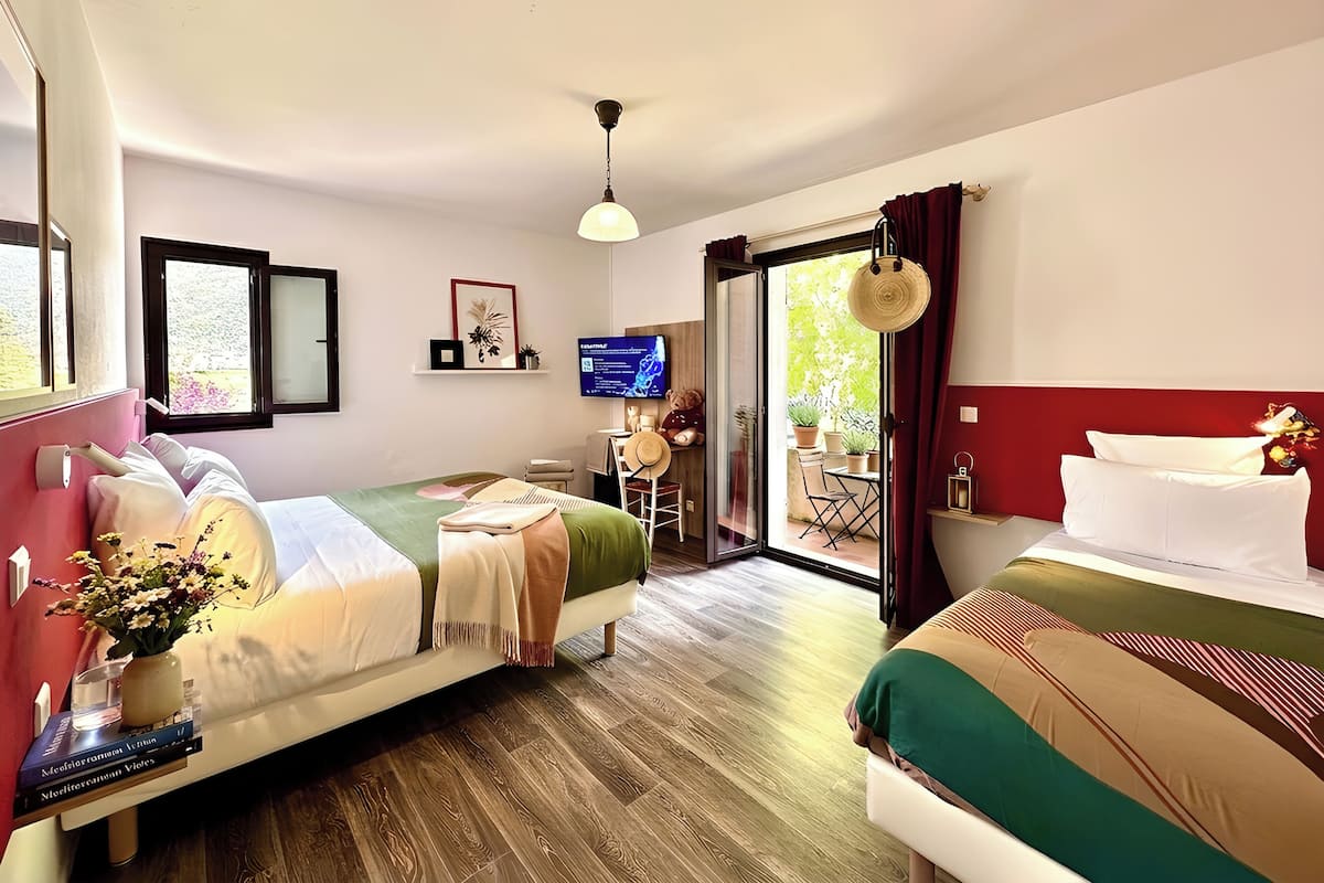 A hotel room with two beds, a TV, a small table with a chair, a window, and a door leading to an outdoor area.