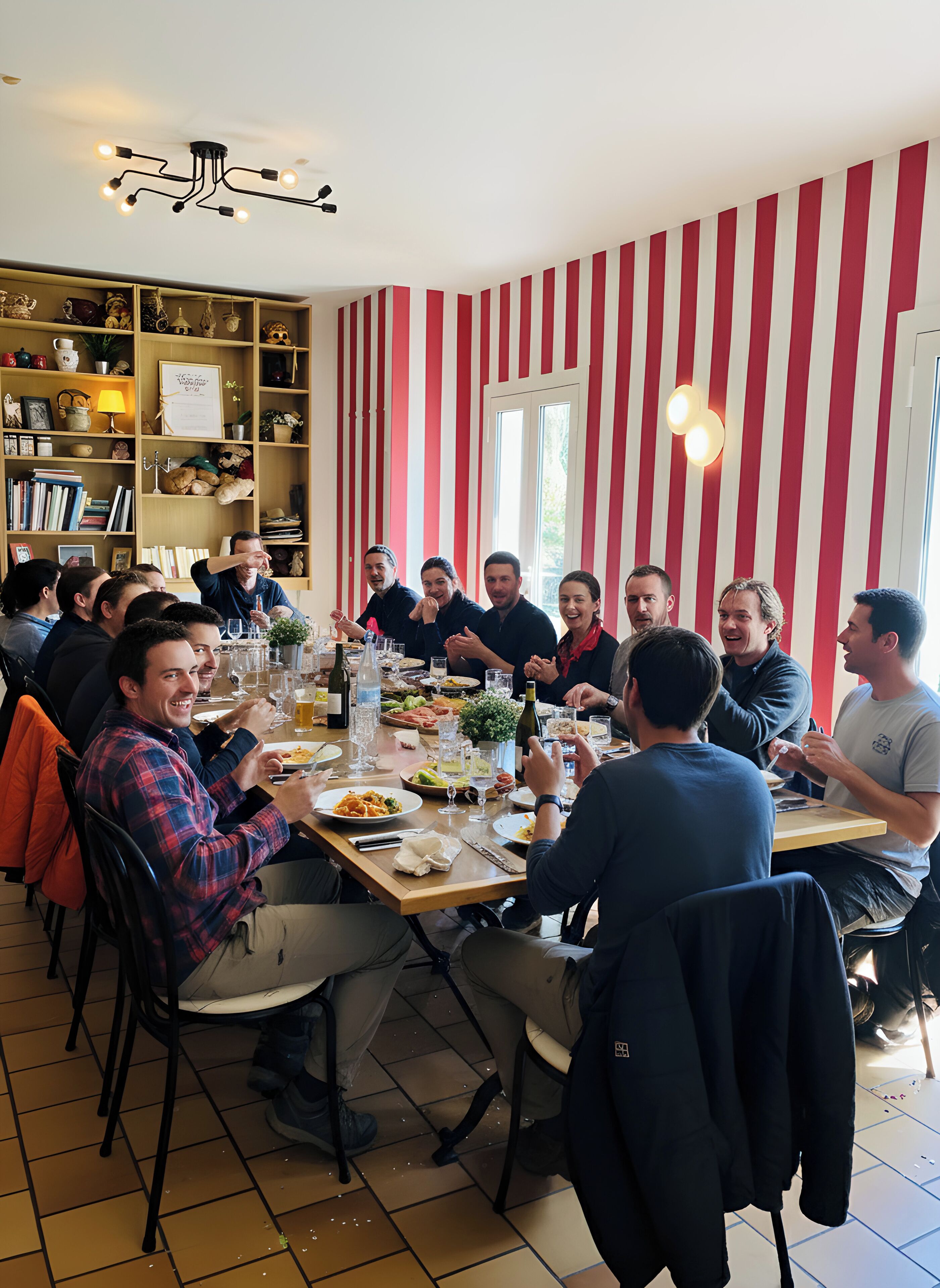 Un grupo de personas cenando en una mesa dentro de una habitación con paredes a rayas y una estantería llena de libros y objetos decorativos.