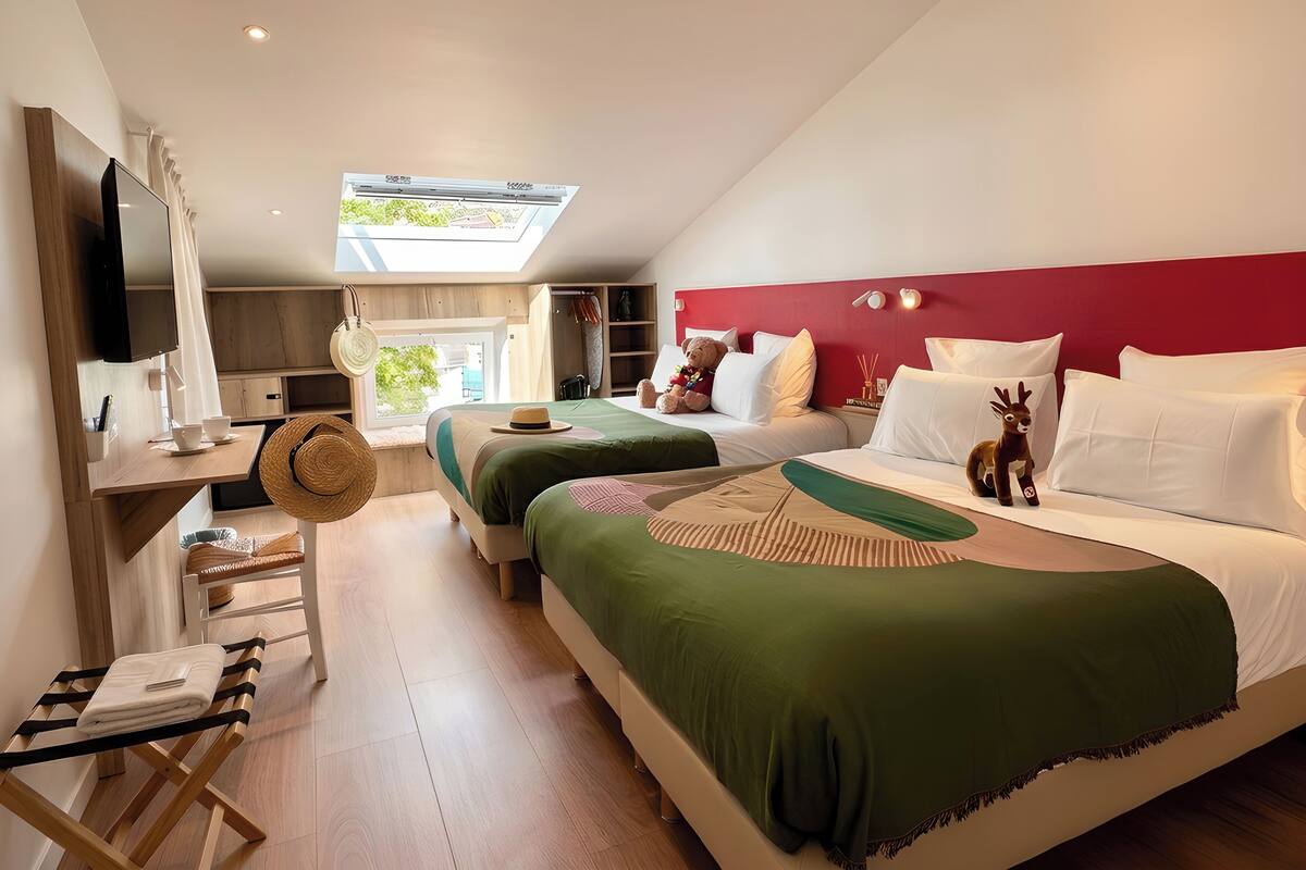 A modern hotel room with two beds, a TV, a desk, and a window with a view of greenery.