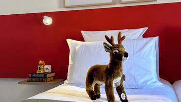 Un lit avec une literie blanche, un ours en peluche, des photos encadrées accrochées au mur et une petite table avec des livres et un objet décoratif.