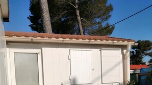 A small, single-story building with a tiled roof, a door, and a window. There is a small table with a potted plant on it, and a chair next to the table. - 2-BEDROOM CHALET IN SUNNY PROVENCE (LE CASTELLET)