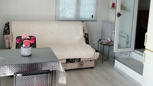 A cozy living room with a sofa, a table, and a chair. There is a window with blinds, a clock on the wall, and a door with a sign. - 2-BEDROOM CHALET IN SUNNY PROVENCE (LE CASTELLET)