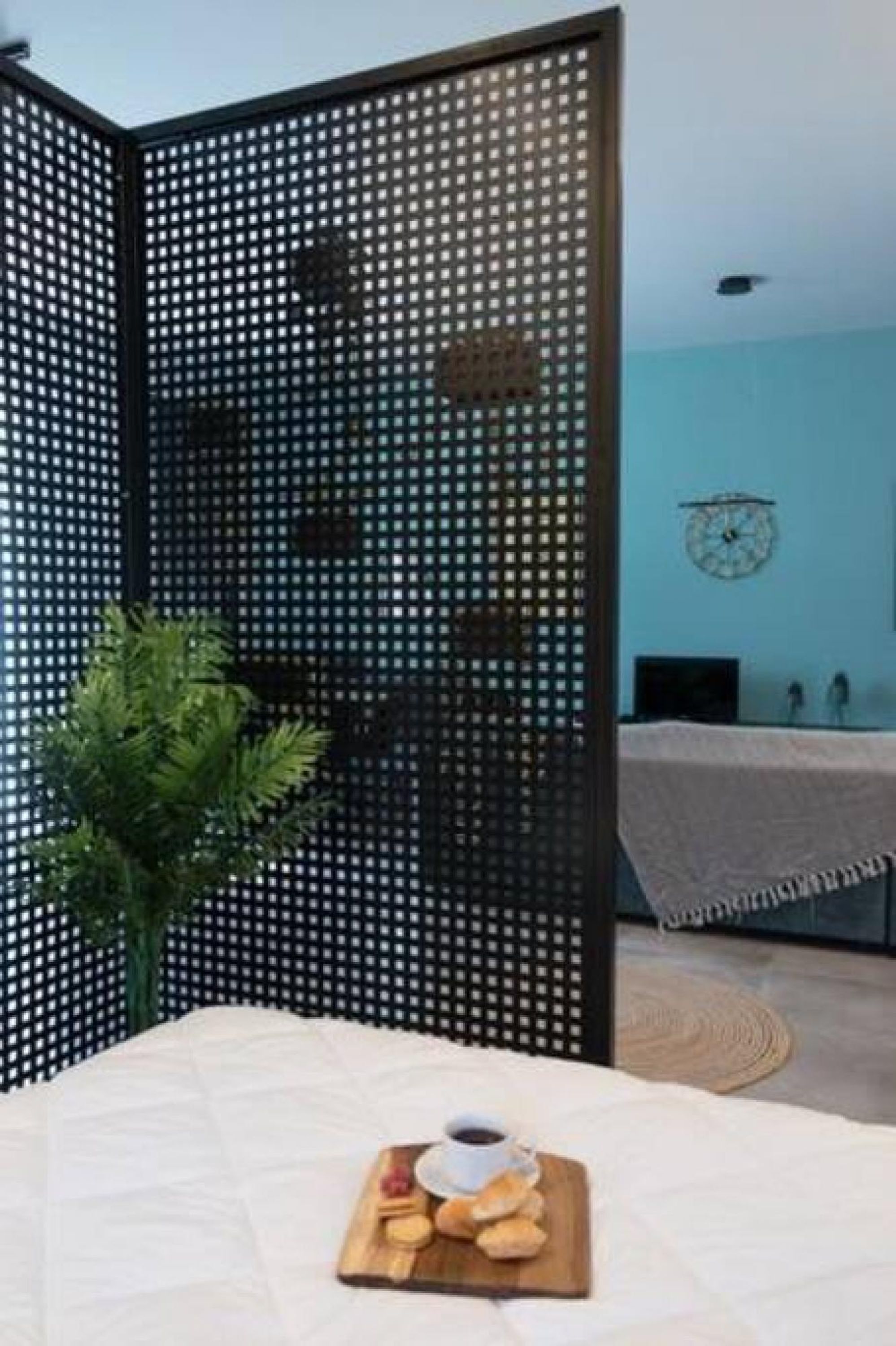 Een kamer met een scheidingswand, een klok aan de muur, een bed met een witte spreidek en een tafel met eten en een kop.