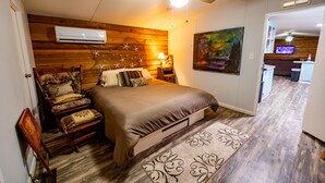Ein Schlafzimmer mit einem Bett, einem Sessel, einer Klimaanlage, einem Deckenventilator, einem Gemälde und einer Küchenzeile im Hintergrund.