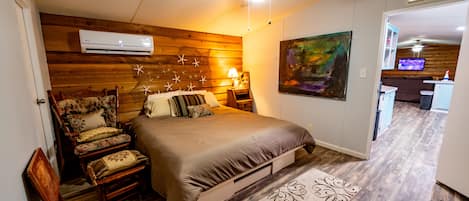 Ein Schlafzimmer mit einem Bett, einem Sessel, einer Klimaanlage, einem Deckenventilator, einem Gemälde und einer Küchenzeile im Hintergrund.