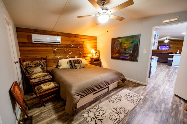 Une chambre avec un lit, une chaise, un climatiseur, un ventilateur de plafond, un tableau et, au fond, une petite cuisine.