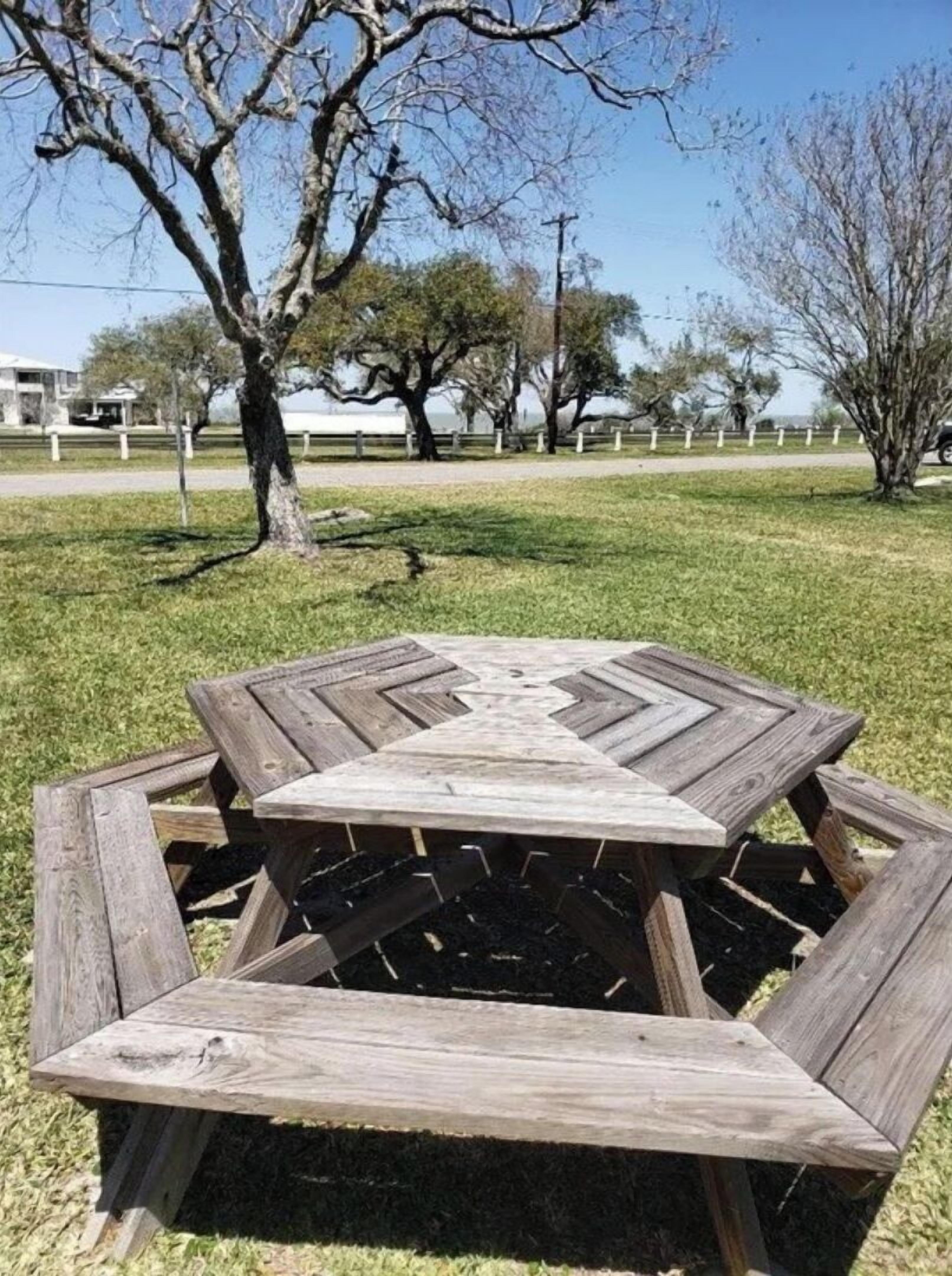 Una mesa de picnic de madera hexagonal con un brasero en el centro, rodeada de césped y árboles en un parque.