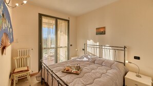 Un dormitorio con una cama, una silla, una mesita de noche, una ventana con cortinas y un cuadro en la pared.