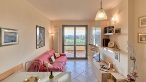 Una sala de estar acogedora con un sofá rosa, una mesa de comedor con una botella de vino y vistas a un paisaje verde a través de puertas de cristal.