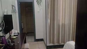 Habitación de hotel con una cama, un escritorio con computadora y monitor, una silla, una mesita con tazas y una planta, y una cortina.