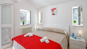 Ein Hotelzimmer mit einem Bett, zwei Fenstern, einem Heizkörper, einem Nachttisch mit Lampe und einer kleinen Topfpflanze.