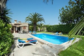 Relax by the serene pool surrounded by lush greenery and palm trees, with lounge chairs and a thatched-roof structure.